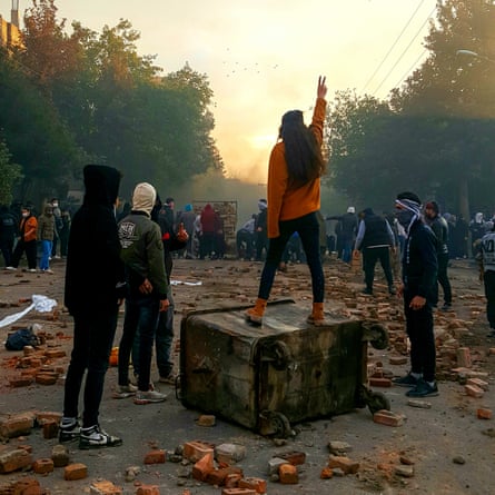 A young woman stands with her back to the camera on a tipped over wheelie bin raising her hand in a ‘v’ sign. Around her are other young protesters wearing hoodies and face scarves. The ground is strewn with broken bricks.