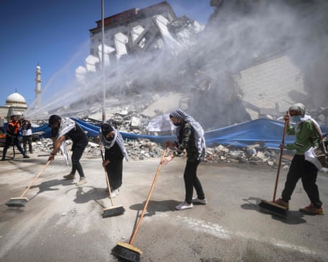 Palestinian volunteers and municipal workers use sweeping brushes on a road, with rubble piled behind