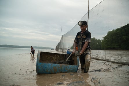 A man knee-deep in muddy water with a head torch on. He has a cut-out blue barrel and he is checking his fishing nets for his catch.