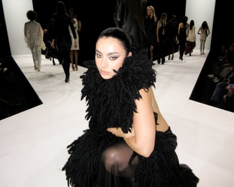 The musician Charli xcx wearing a black feather boa while crouched on a white X-shaped catwalk as models parade in the background