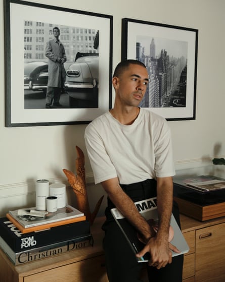 A man in a white t-shirt leaning against a side cabinet, near a wall with two black and white photographs.