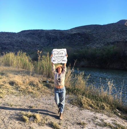 A woman stands near a river and holds up a sign that says ‘Border Cultura’ inside a heart and ‘No Wall’ underneath