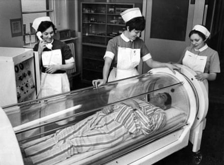 Three nurses look over a man who is resting in a see-through chamber