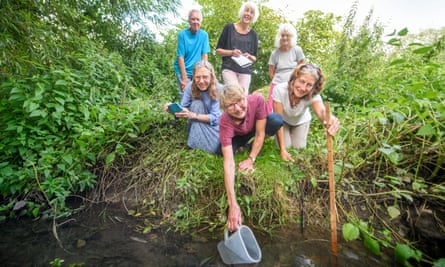 Five older women and one man on the edge of the River Wye, one dipping a plastic jug into the water