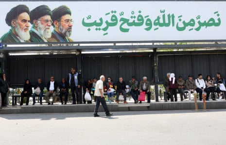 Over a dozen people wait at a bus stop displaying a poster of Iran's current and former supreme leaders.