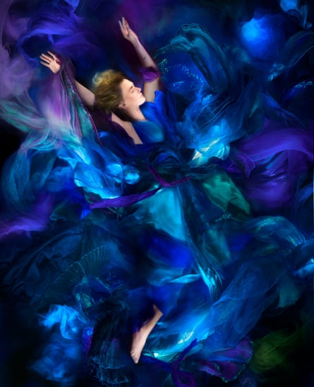 A woman with her eyes closed and arms outstretched surrounded by billowing cloth in different shades of blue, purple and indigo