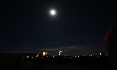 A mostly black image, with a bright white circle of light in the sky, and what you discern is a circle on the ground of hot air baskets, with each one shooting flames skyward.
