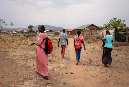Four people walk along a red dirt road towards some low-rise buildings