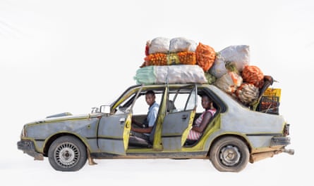 Taxi driver Cheikh and market vendor Arame in a Renault in Noto, Senegal. Cheikh is in the front seat Arame in the back of the grey/silver car with both doors open and all her produced stacked on the roof and boot.