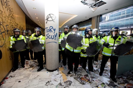 Police respond to the riot in Millbank Tower, with riot shields. A pillar is graffitied with an anarchist symbol and “Tory scum”