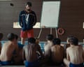 A coach stands and talks to a group of boys who sit on the floor in front of him at a swimming pool.