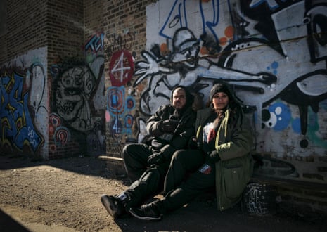 Two men sit on a bench in front of a graffiti covered wall.