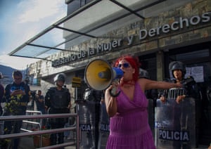 A woman in a bright pink dress speaks into a loudspeaker as police officers with riot shields stand behind her
