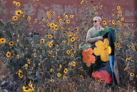 Andy Warhol in a field of flowers holding one of his flower paintings, against a wall with peeling paint