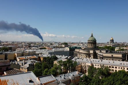 A dark cloud of smoke billows in the distance over the city. The view shows historic buildings and churches, and the smoke is thick in a clear blue sky.