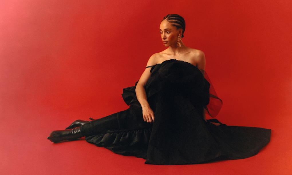 Adwoa Aboah sitting down wearing a full black dress and black boots, on a red backdrop