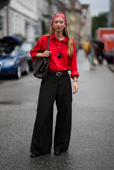Woman with long blond hair, standing on a street wearing a red shirt, black wide trousers and a white and red checkered bandana