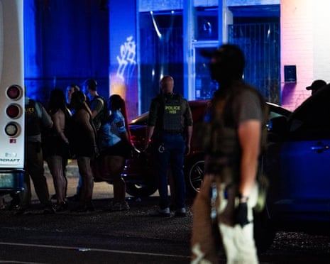 Woman in miniskirts lined up in handcuffs behind their backs to be loaded into van by police, at night with blue light on wall of building.