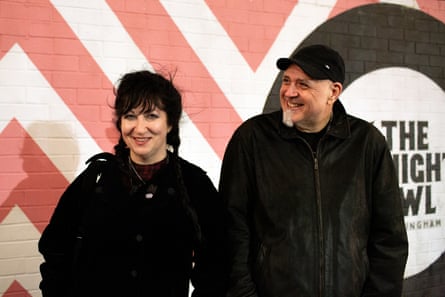 A man in a black jacket and black cap, and a woman in a black jacket, in front of a red and white striped wall, and a painted disc saying ‘The Night Owl’.