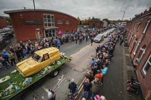 A funeral cortege, including a Reliant Robin car on a trailer, with people standing and watching from the street