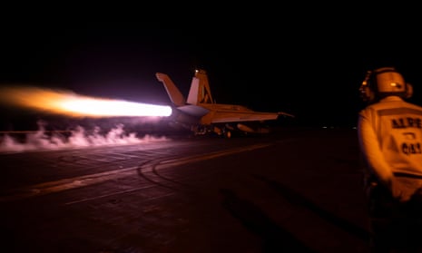 A fighter jet takes off from an aircraft carrier at night as a crew member looks on.
