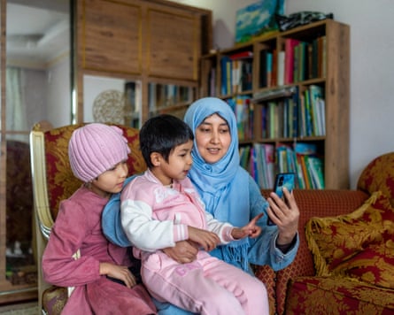 A woman and two children sit inside looking at a phone. Furniture including a bookcase can be seen behind them