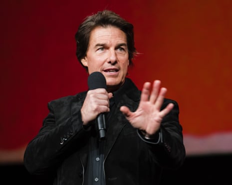 Tom Cruise, wearing all black, speaking into a microphone against a red background