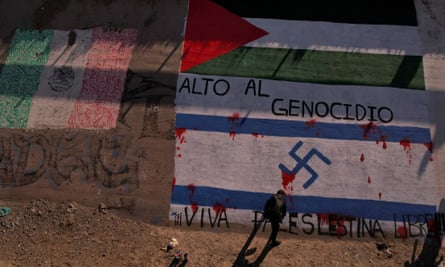 a multistory concrete barrier is covered with paintings of palestinian and israeli flags, with what appears to be a young man dressed in black, his shadow long behind him on the sign, painting the bottom, israeli flag