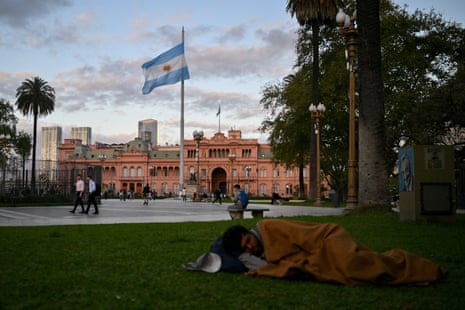 poverty rate - A man sleeps on the ground in front of a government palace with a large Argentinian flag waving in the wind.