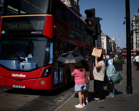 People use umbrellas and shopping bags to shield themselves from the sun while waiting to cross Oxford Street.