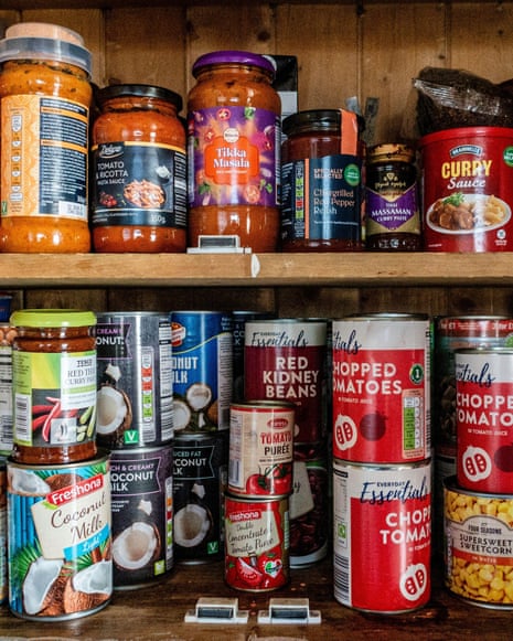 Wooden pantry shelves hold jars of curry sauce and many tinned goods like tomatoes and beans