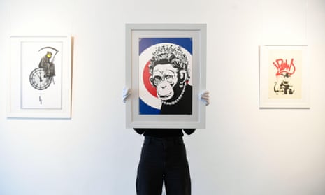 A person with gloves blocks their face and chest by holding up the framed Banksy print Monkey Queen, which shows a monkey wearing a necklace and crown, with the Banksy prints Grin Reaper and Gangsta Rat on the wall behind