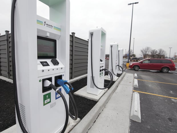 An electric car charging station is seen in a parking lot. A few spaces down is a red gas-powered minivan.