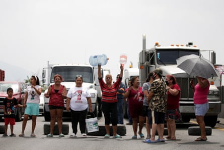Women and a child wave empty water bottles as they block a highway with trucks behind them 
