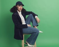 A woman sitting on a stool, wearing a white turtleneck, navy jacket, blue flare jeans and white slingback shoes