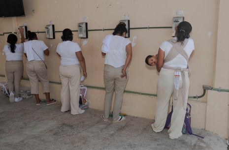 Five women stand in a row, one holding a baby, as they make calls with wall-mounted phones at the prison