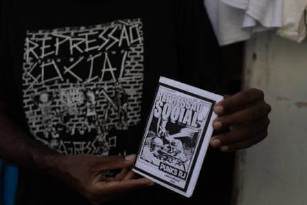A person wearing a black T-shirt with bold white text holds a small booklet titled ‘Repressão Social – Punks RJ,’ featuring a black-and-white illustration on the cover.