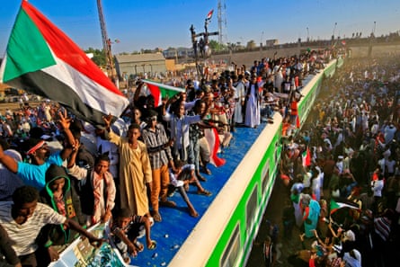 Büyük bir kalabalık trenin etrafında duruyor ve çatıda Sudan bayrakları sallıyor