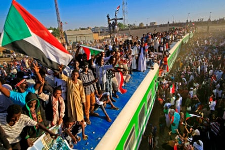 A large crowd of people stand around a train and on its roof waving Sudanese flags