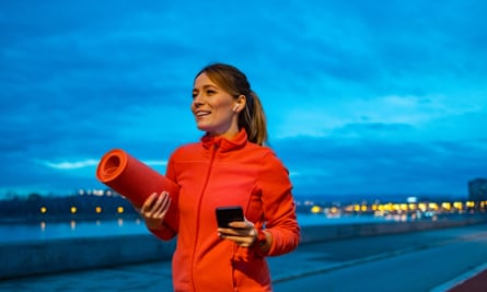 A woman in gym kit, holding a yoga mat, listening to music while walking outdoors