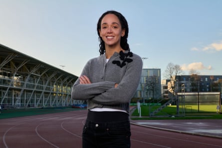 A young woman wearing a grey cardi and black trousers standing with arms folded with an athletic stadium behind her