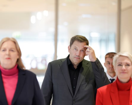 Lars Klingbeil in a grey suit scratching his forehead walking between Baerbel Bas and Manuela Schwesig, respectively wearing pink and red turtleneck jerseys