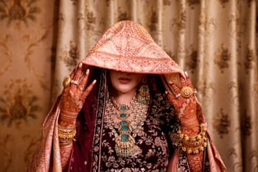 A woman holds a scarf over her head and part of her face. She has henna on her arms and hands and elaborate jewellery including a necklace, bangles and rings