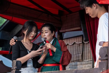 Granny Wang puts her arm around a young woman on stage during her show as she speaks into a microphone; a tall young man in a white T-shirt looks on, smiling