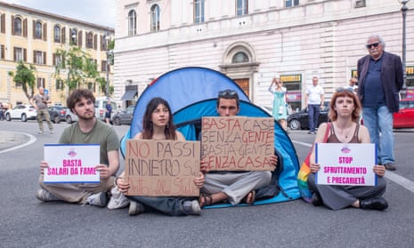 Four students sit on the road in front of a blue tent, holding signs protesting against high rents.