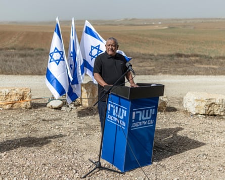An older man stands by a blue podium on a big field with three Israeli flags behind him
