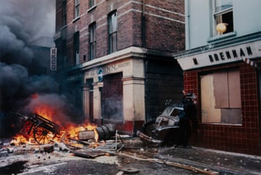 In Derry, a police officer stands near a burning cart and wrecked car outside brick buildings