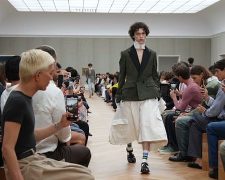 A male model walks down the catwalk wearing a fitted mossy green dinner jacket with black velvet collar, a white bow tie but no shirt, and huge baggy cream below-the-knee shorts, plus white socks and sandals