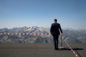 A rear shot of a person in a dark blue suit standing next to red and white cordon tape while holding on to a silver post and looking at mountain peaks on a clear day
