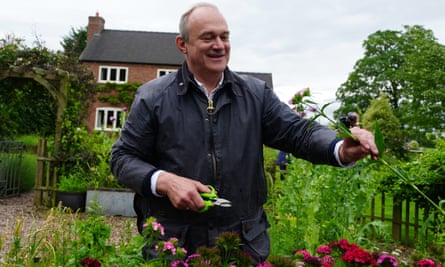 Ed Davey picks flowers at Shropshire Flower Farm while on the general election campaign trail: he is seen with a pair of secateurs, leaning towards a tall flower, with a garden, trees and a house behind him. He wears a waxed jacket and is smiling.